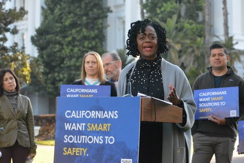 black woman outdoors at lectern with sign that reads "californians want smart solutions to safety." Other people, including a man holding the same sign, and tall greenery obscuring a white building are behind her