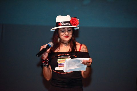 A Latina woman wearing a white hat that says Xicana on a black band, a red rose on the hat, and a black shirt that says La Raza studies contra costa college with pictures of books. She is holding a microphone and piece of paper