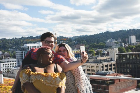 Three smiling and fashionably dressed heavyset people taking a selfie on what appears to be the roof of a building with other buildings and tree-covered hills in the background