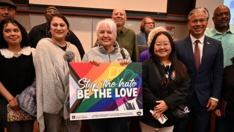 A group of people including the california attorney general standing together with one older white woman holding a sign decorated with the progress pride flag and a raised fist and text that reads stop the hate be love