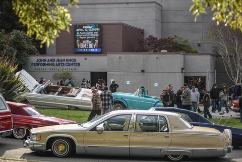 Lowrider cars and people in front of the John and Jean Knox performing arts center at contra costa college. electronic sign on the building reads friday april 12 5:30 p.m. la raza studies and automotive collision repair present american homeboy ticket required