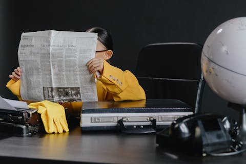 Little girl at a big desk reading the new york times. the desk is holding a globe, briefcase, corded phone and yellow rubber gloves