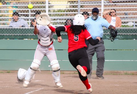 A softball player and the ball are both approaching home plate with the catcher standing up and umpire looking on
