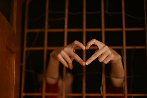 A person's hands sticking through wire bars making the shape of a heart with their index and middle fingers. The person is mostly in shadow inside the cell.