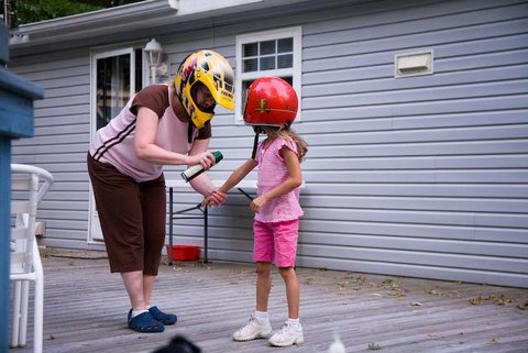 a woman spraying bug spray on a girl's arm. they are standing outside a house and both wearing motorcycle style helmets