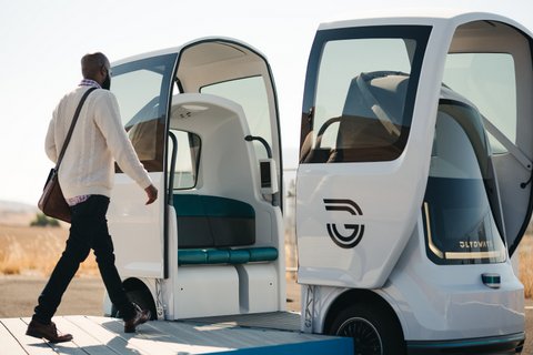 A black man seen from the side walking on a platform toward a small futuristic looking vehicle. Rather than having traditional doors, the top and sides in the middle move to open the car.