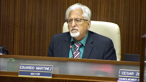 An older Latino man white hair and goatee in suit seated with microphone and nameplate identifying him as eduardo martinez mayor in front of him