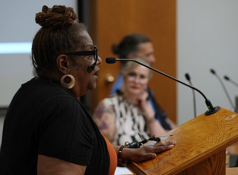 Side view of a black woman standing at a lectern with a seated white woman looking at her from the other side and an out of focus man sitting past the white woman