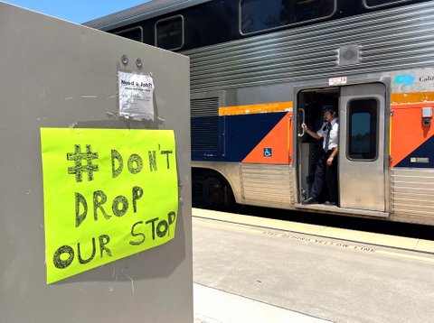 A sign that reads don't drop our stop with a hashtag symbol taped near a stopped amtrak train with a worker standing in the open door