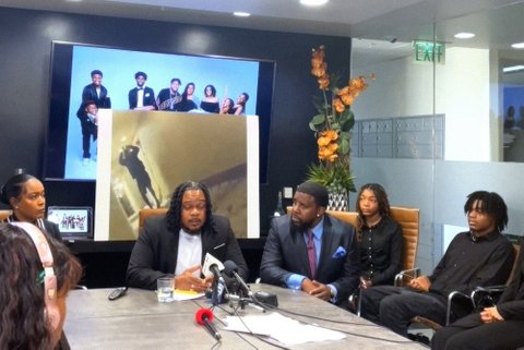 a black man sitting at a conference table in front of microphones with a black man in suit and tie and a black woman on either side of him. also seated in the room are a black boy and girl who appear to be in their teens
