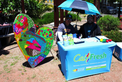 Two men sitting under an umbrella with two alternating shades of blue on a sunny day. The table is draped with a light blue banner that says "Cal Fresh," "Food Bank" and "grocery assistance" and "apply now" in english an spanish. next to them is a large colorfully painted heart figurine