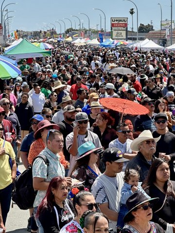large crowd of people on city street. people visible in the foreground are hispanic. some people have umbrellas or little mexican flags. tent top stands are set up, and a tall u haul sign is visible