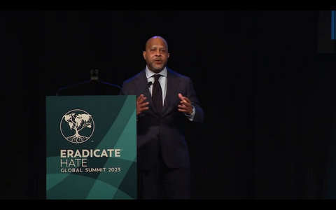 A black man in a suit standing next to a lectern labeled eradicate hate global summit 2023
