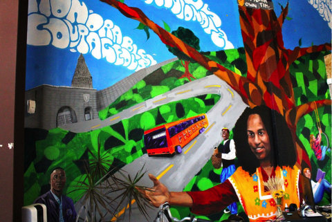a colorful mural that includes illustrations of a smiling black man with an arm extended, bus going down a street, greenery and a tree with a person reading a book under it and a child on a tire swing