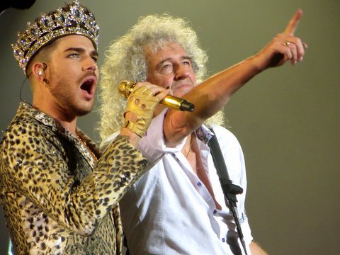 adam lambert wearing a leopard print suit and crown singing into a golden microphone next to brian may who has long curly gray hair and is pointing