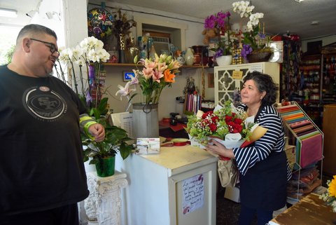 a latina woman wearing a black and white striped shirt holding a bouquet of red roses standing across from a taller, heavyset latino man in a small shop with other flowers around