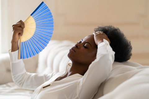 a black woman dressed in white sitting on a white couch with her head back, eyes closed and hand on her forehead as she fans herself with a blue and yellow paper fan