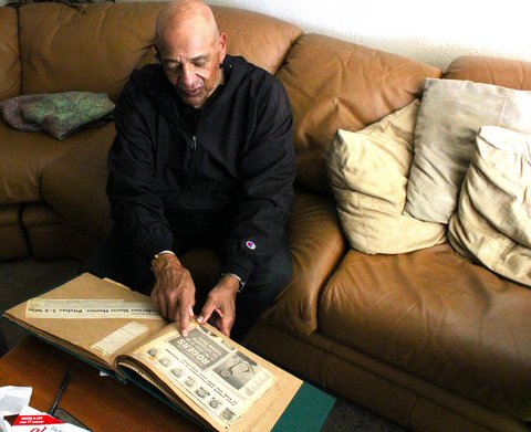an older bald black man pointing to a newspaper clipping in a scrapbook he is holding while sitting on a brown couch