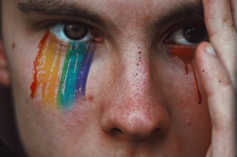 closeup of a white person with a serious expression, one hand to their face, rainbow colors painted from the bottom of one eye to their cheekbone and a drop of red streaming from the other eye