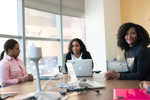 Three black women at a conference table with two laptops open. one woman is smiling at the camera