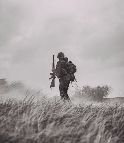 Grayscale photo of a soldier seen from behind holding a rifle against a hazy sky