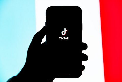 A hand shrouded in darkness holding up a smartphone with the tik tok logo on the screen. this is in front of a light blue, white and red background
