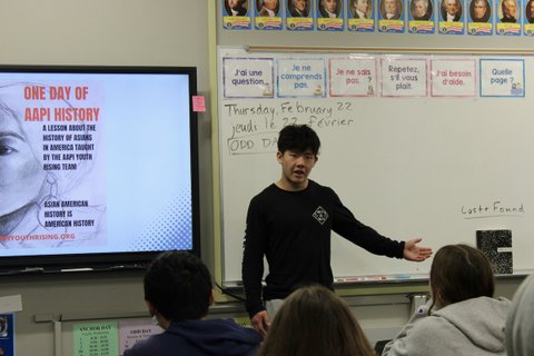 an asian man standing in front of a classroom. a screen behind him says one day of AAPI history. a lesson about the history of asians in america taught by the AAPI youth rising team. asian american history is american history.