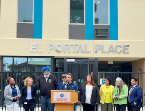 a white man standing at a lectern that says contra costa health on it. in a row behind him are a black woman, white woman, tall indian man with gray beard and blue turban, man in pork pie hat and dark sunglasses, three other women and a man in a suit. All are standing in front of a building that says el portal place