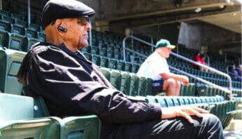 side view of an older black man sitting in a stadium. he is dressed in black and wearing a black flat cap, dark sunglasses and a bluetooth earpiece
