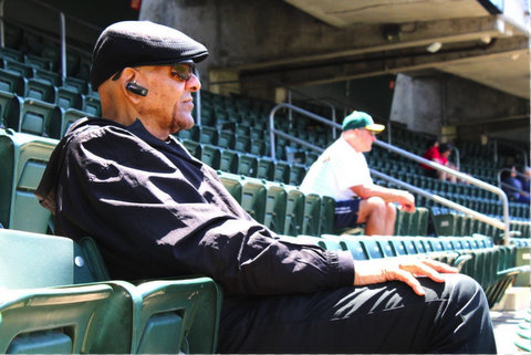 side view of an older black man sitting in a stadium. he is dressed in black and wearing a black flat cap, dark sunglasses and a bluetooth earpiece