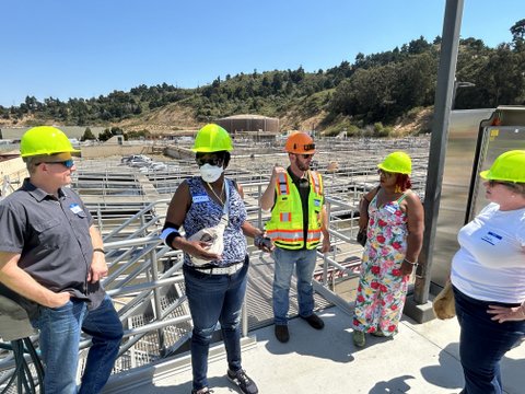 two white men, two black women and a white women all wearing hard hats and standing outdoors on a sunny day at a water treatment facility