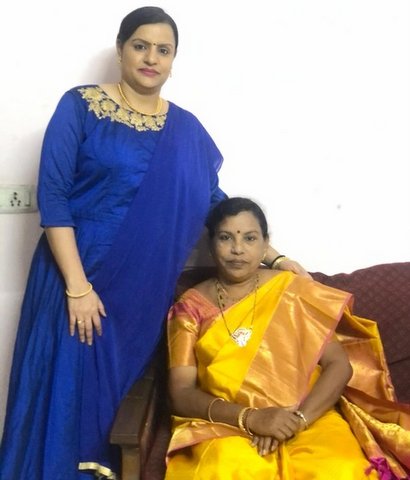 two indian women in traditional dress. one is wearing blue and standing next to the other who is seated on a couch and dressed in a gold color