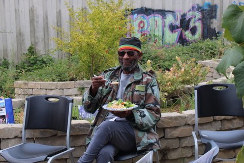 black man with graying beard, sunglasses, black kufi hat with green, red and yellow stripes. He is sitting with a plate of food in front of greenery