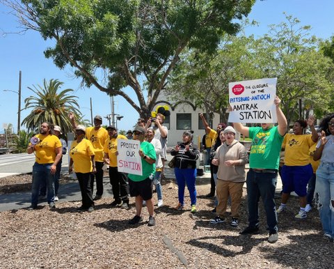 A group of people including one holding a sign that reads protect our station and another with a sign that reads stop the closure of the antioch pittsburg amtrak station with stop depicted like a stop sign