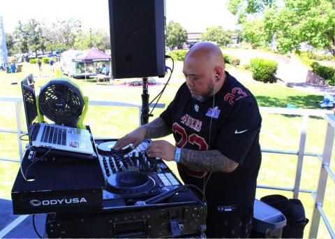 man with shaved head, beard, black 49ers jersey, tattoo sleeve on left arm stands at a turntable next to a loudspeaker with a laptop that has a fan pointed at it