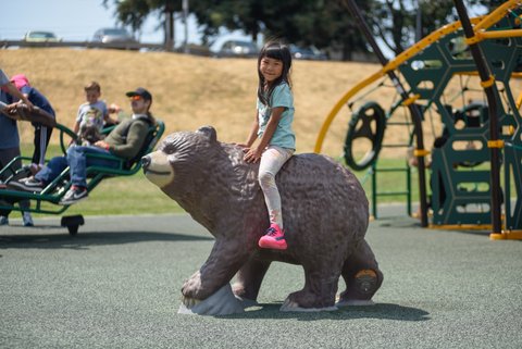 a little asian girl sits straddling a bear statue on a playground