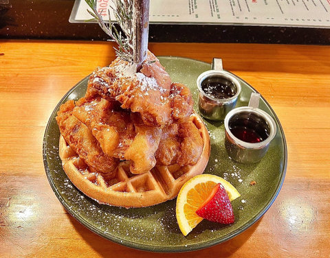 A green plate with a waffle with fried chicken on top of it and a knife and rosemary sprigs sticking vertically out of it. Also on the plate are two containers of syrup and an orange and strawberry slice