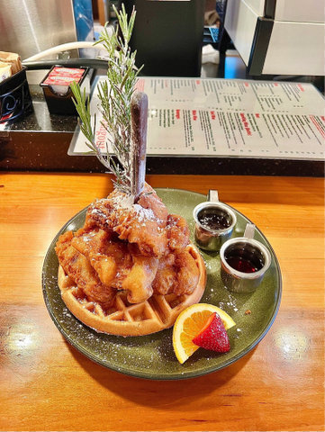 A green plate with a waffle with fried chicken on top of it and a knife and rosemary sprigs sticking vertically out of it. Also on the plate are two containers of syrup and an orange and strawberry slice. visible beyond the plate are a laminated menu and a container of smuckers jelly packets