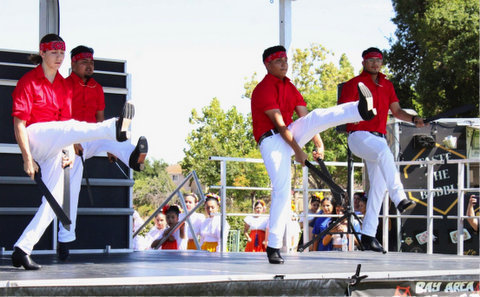 four male dancers on stage wearing a red bandanna worn around their foreheads like a headband, red shirts and white pants. each one has a machete in each hand, one leg off the ground and their other heel slightly raised