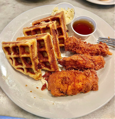 plate with four triangular waffle slices, three pieces of fried chicken and plastic cups of butter and syrup. the tines of a fork are resting on the edge