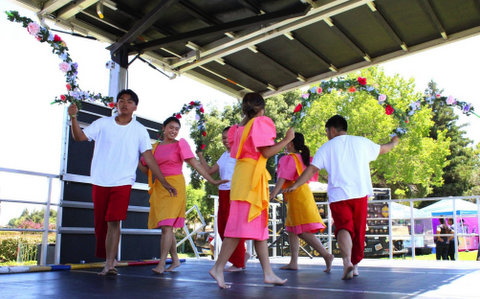 barefoot filipino dancers in a ring on stage. the male dancers are wearing white t shirts and red pants with one leg rolled up to the knee. the female dancers are wearing pink and yellow dresses. all are each holding a curved flowered branch of sorts