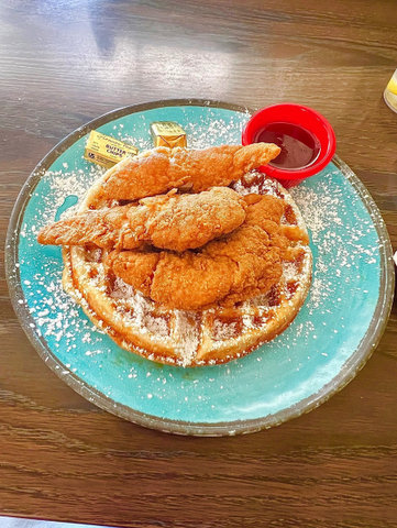 a blue plate with waffle topped with fried chicken, generously dusted with powdered sugar, two gold foil wrapped pats of butter and a little red ramekin of syrup