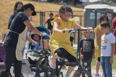 a little girl in yellow dress, black shorts and glasses pedals a stationary bike with a blender attached to the back that a woman wearing an apron and gloves is operating