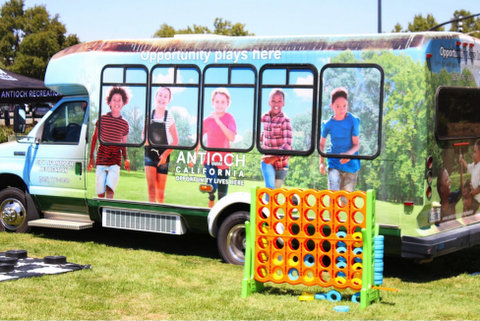 bus parked on grass. a photo of five kids running in a park covers one side. two black adults and a child are in the picture on the back. text on the bus reads "opportunity lives here" multiple times and "antioch california." next to the bus is a large connect four style game and the corner of a life size checkerboard is visible