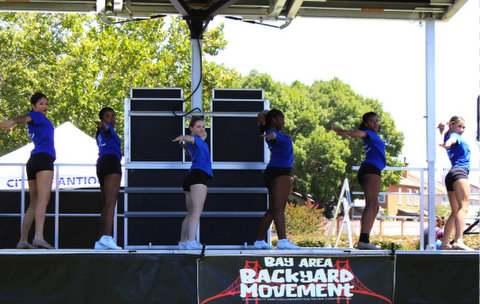 six young women in blue shirts and black shorts on stage with their bodies facing the viewer's right but mostly looking toward the audience (not pictured). each has their right arm extended out toward the audience and their left hand curled toward their head