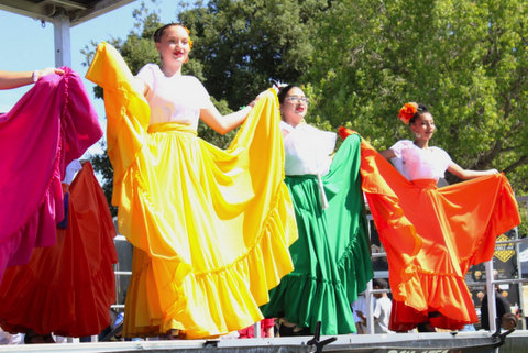 three latina young folklorico dancers on stage holding their skirts spread out to the sides. their skirts are, from left, yellow, green and orange. another dancer's magenta skirt is visible at far left