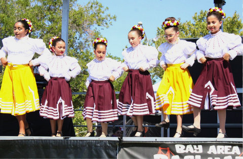 latina girls wearing white peasant blouses and skirts with their hair in buns adorned with a braided crown. two are wearing yellow skirts and four are wearing maroon. the skirts have four white stripes at the bottom