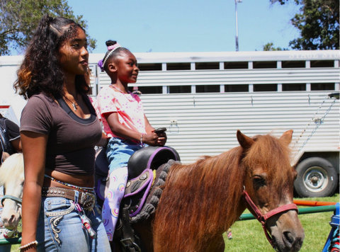 a little black girl riding a pony with a black teen girl standing next to a her and a pony's head visible behind her