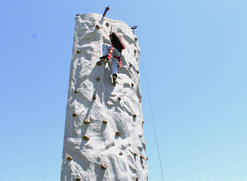 a tall outdoor fake rock wall against a blue sky with one climber on it