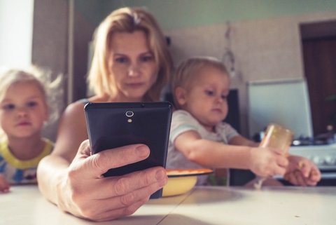 a white woman looking at a smartphone with a baby on one side of her and a small child on the other